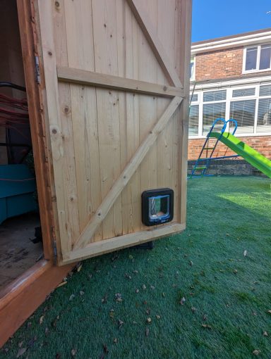 Wooden shed door with a cat flap, set on lawn near a playground slide.